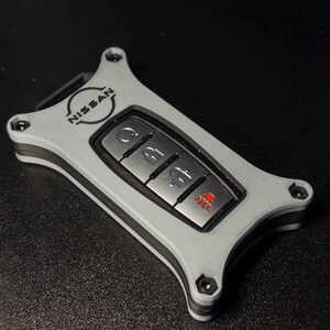 Può includere: Una chiave elettronica Nissan inserita in un guscio protettivo grigio chiaro a forma di clessidra, fissato con quattro viti nere agli angoli. Il telecomando metallico presenta quattro pulsanti con icone per blocco, sblocco e allarme rosso.