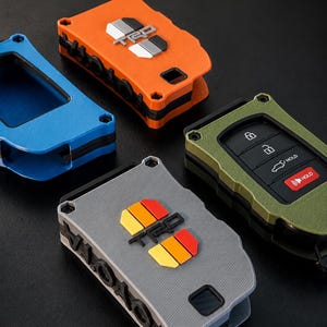 May include: Four rectangular key fob covers in orange, blue, olive green, and gray. The orange and gray covers feature the TRD logo with colored accents. The olive green cover displays a black key fob with lock, unlock, and hold buttons.