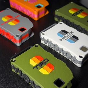 May include: A collection of five rectangular key fob covers in orange, gray, white, and olive green. Each cover features embossed Toyota branding and TRD or Hilux logos with colorful striped accents. The covers have visible screw holes and cutouts.