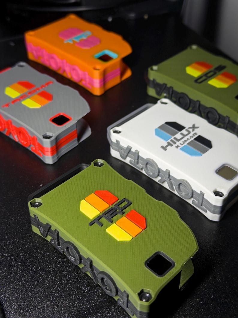 May include: A collection of five rectangular key fob covers in orange, gray, white, and olive green. Each cover features embossed Toyota branding and TRD or Hilux logos with colorful striped accents. The covers have visible screw holes and cutouts.