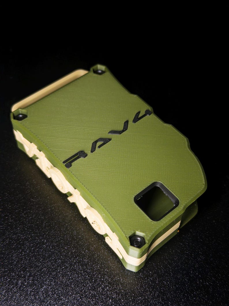 May include: A rectangular olive green phone case with a tan inner layer and black accents. The word RAVE is embossed in black on the back surface. The case features a square camera cutout and a textured finish with visible 3D printed layer lines.