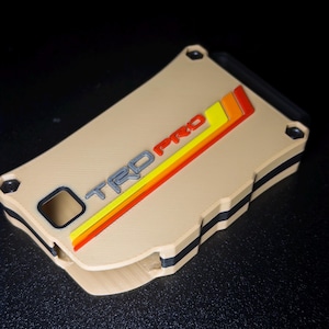 May include: A rectangular tan wallet with a black interior layer and four corner screws. The front features the embossed text TRD PRO in grey, accompanied by three diagonal stripes in yellow, orange, and red, all set against a solid tan surface.