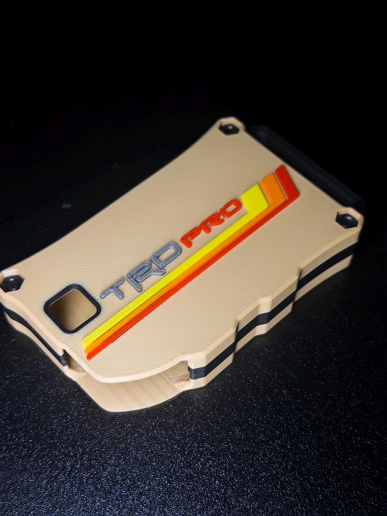 May include: A rectangular tan wallet with a black interior layer and four corner screws. The front features the embossed text TRD PRO in grey, accompanied by three diagonal stripes in yellow, orange, and red, all set against a solid tan surface.