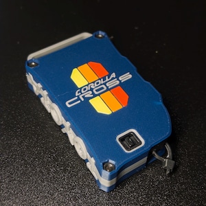 May include: A rectangular blue key fob cover featuring the text COROLLA CROSS in white lettering. The design includes a vertical graphic with yellow, orange, and red stripes. The case has a grey side frame, visible metal screws, and a small black loop.