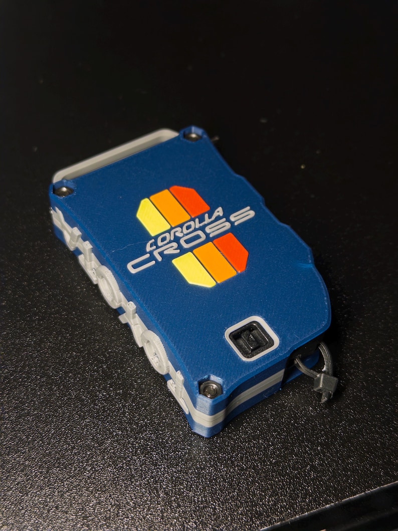 May include: A rectangular blue key fob cover featuring the text COROLLA CROSS in white lettering. The design includes a vertical graphic with yellow, orange, and red stripes. The case has a grey side frame, visible metal screws, and a small black loop.