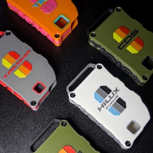 May include: Five rectangular key fob covers in orange, olive green, gray, and white. The covers feature embossed logos including TRD, Tacoma, and Hilux Kumar with colorful striped accents. Each case has a rugged design with visible screw details.
