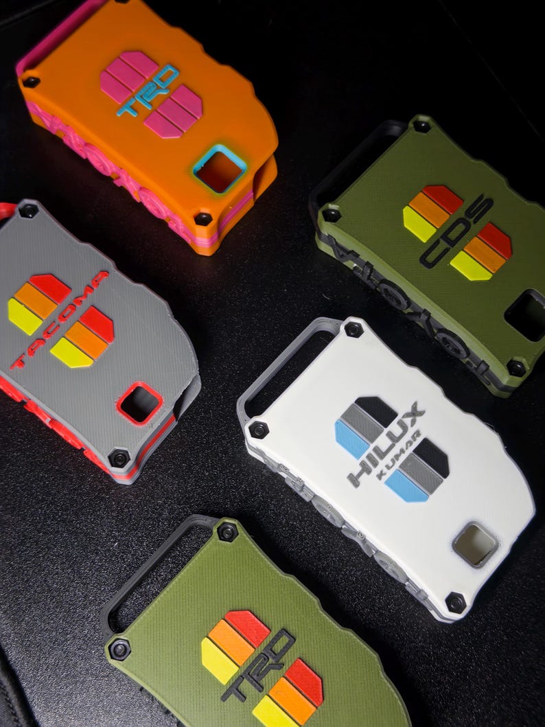 May include: Five rectangular key fob covers in orange, olive green, gray, and white. The covers feature embossed logos including TRD, Tacoma, and Hilux Kumar with colorful striped accents. Each case has a rugged design with visible screw details.