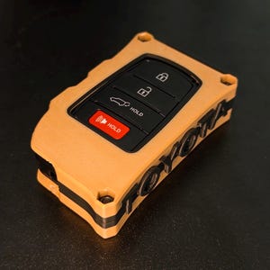 May include: A tan and black key fob cover featuring a rectangular shape with a cutout for the black control buttons. The side of the cover displays the word TOYOTA in raised black lettering. The buttons include lock, unlock, and hold icons.