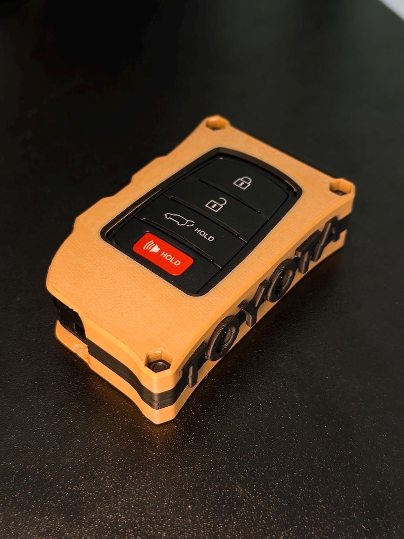May include: A tan and black key fob cover featuring a rectangular shape with a cutout for the black control buttons. The side of the cover displays the word TOYOTA in raised black lettering. The buttons include lock, unlock, and hold icons.