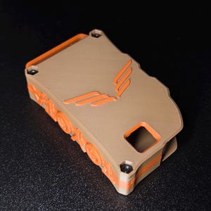 May include: A rectangular, 3D-printed case featuring a tan top panel with an embossed orange wing-like logo. The sides are orange with circular gear-like details and a square cutout. Four small black screws secure the tan top to the orange base.