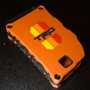 May include: An orange rectangular key fob cover featuring the black text TRD centered above a graphic of two yellow and red stripes. The sides of the cover are black with a textured pattern of small circular and geometric shapes along the edges.