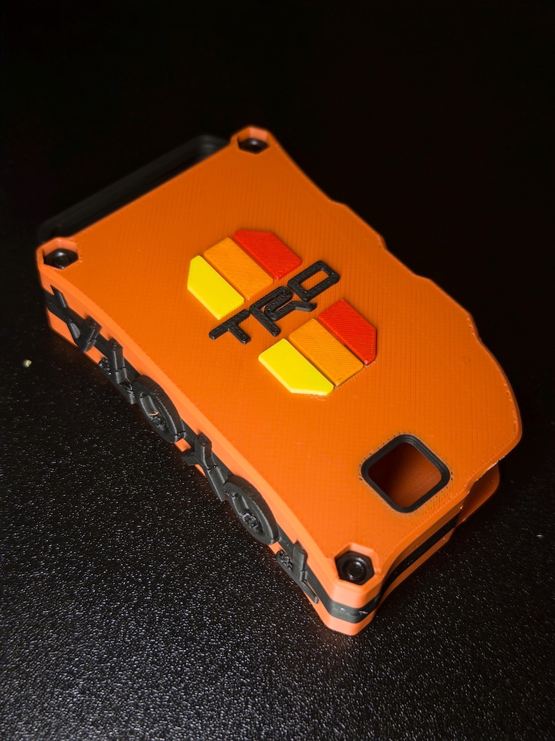 May include: An orange rectangular key fob cover featuring the black text TRD centered above a graphic of two yellow and red stripes. The sides of the cover are black with a textured pattern of small circular and geometric shapes along the edges.