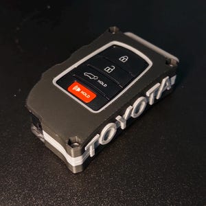 May include: A black and gray protective key fob cover for a Toyota vehicle. The cover features a textured finish with the word TOYOTA embossed in white lettering on the side. The internal key fob has black buttons with white lock, unlock, and trunk icons, plus a red button labeled HOLD.