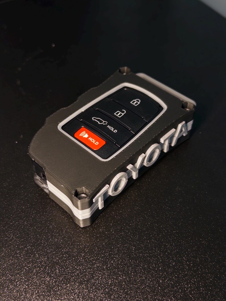 May include: A black and gray protective key fob cover for a Toyota vehicle. The cover features a textured finish with the word TOYOTA embossed in white lettering on the side. The internal key fob has black buttons with white lock, unlock, and trunk icons, plus a red button labeled HOLD.