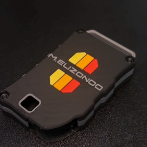 May include: A rectangular black key fob case with a layered design featuring grey accents and four screws at the corners. The text M.ELIZONDO is printed in white, accompanied by two sets of three small, vertical yellow, orange, and red rectangles.
