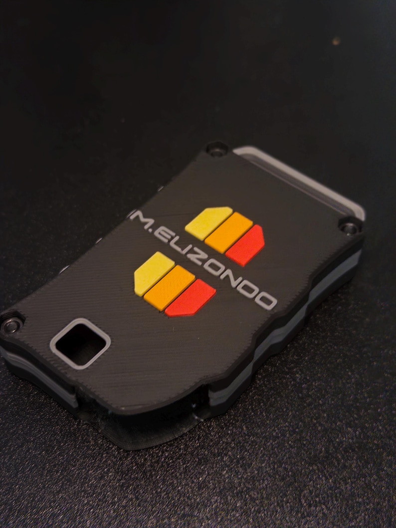 May include: A rectangular black key fob case with a layered design featuring grey accents and four screws at the corners. The text M.ELIZONDO is printed in white, accompanied by two sets of three small, vertical yellow, orange, and red rectangles.