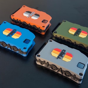 May include: Four rectangular key fob covers in orange, blue, olive green, and gray. Each cover features the word TOYOTA embossed on the side and a TRD logo with a horizontal three-stripe graphic in yellow, orange, and red on the top surface.