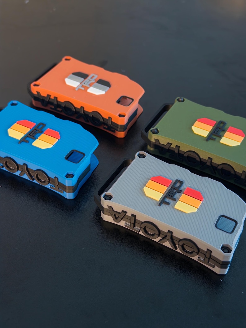 May include: Four rectangular key fob covers in orange, blue, olive green, and gray. Each cover features the word TOYOTA embossed on the side and a TRD logo with a horizontal three-stripe graphic in yellow, orange, and red on the top surface.