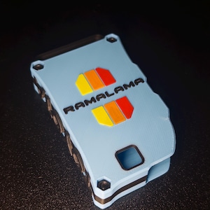 May include: A light blue rectangular case with black accents and a textured surface. The word RAMALAMA is printed in black, flanked by two sets of yellow, orange, and red rectangular shapes. The case features visible black screws at the corners.
