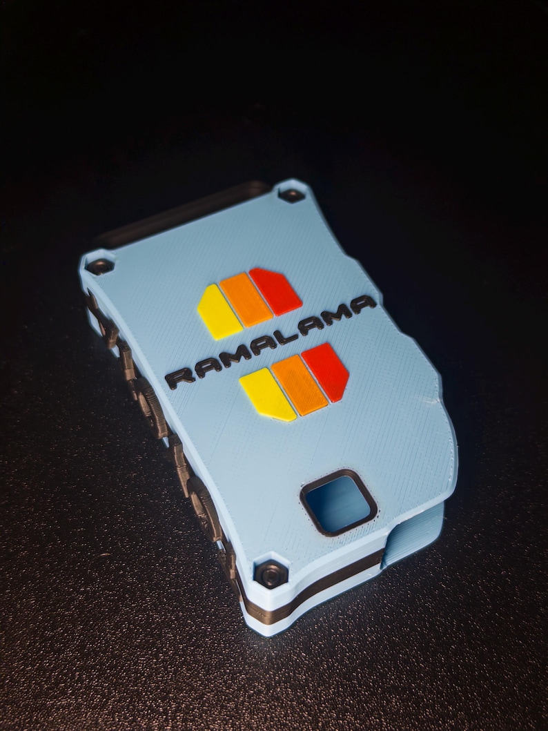 May include: A light blue rectangular case with black accents and a textured surface. The word RAMALAMA is printed in black, flanked by two sets of yellow, orange, and red rectangular shapes. The case features visible black screws at the corners.
