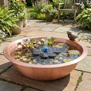 May include: A round copper-colored basin filled with water, smooth river stones, and green aquatic plants. A black flower-shaped solar fountain pump sits in the center, spraying a vertical stream of water. A small bronze bird figure sits on the rim.