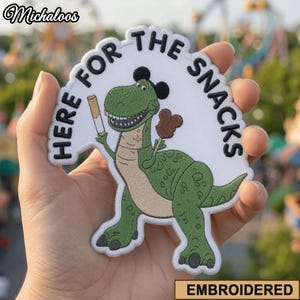 Pode incluir: Um emblema bordado com um dinossauro verde usando orelhas de rato pretas, segurando um churro e um snack castanho em forma de rato. O emblema branco tem o texto preto HERE FOR THE SNACKS, com uma borda branca e formato recortado.