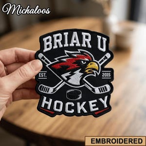 May include: A black embroidered patch featuring the text BRIAR U HOCKEY in white, with a central graphic of a red, white, and yellow eagle head mascot positioned over two crossed white hockey sticks and a black hockey puck, with EST. 2015.