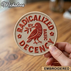 May include: A circular embroidered patch with a beige background and rust-coloured stitching. The text reads RADICALIZED BY BASIC DECENCY, surrounding a central bird illustration holding a small branch. Two small flower icons flank the bird.