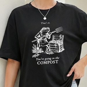 Op de afbeelding: Zwart t-shirt met een witte afbeelding van een kikker met een hooivork en een schatkist. De tekst luidt "That's it." boven de kikker en "You're going in the COMPOST" onder de afbeelding.