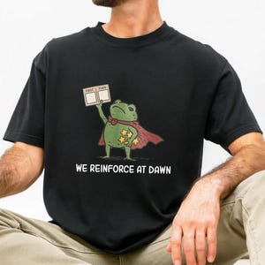 Peut inclure: Un t-shirt noir avec un motif représentant une grenouille verte portant une cape rouge, tenant un panneau indiquant FIRST THEN et un groupe d'étoiles jaunes. Sous la grenouille, le texte WE REINFORCE AT DAWN est imprimé en lettres blanches.