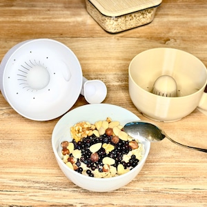 Può includere: Uno spremiagrumi bianco con la parte superiore rotonda e un beccuccio, accanto a una ciotola di frutta secca e bacche miste. Un cucchiaio in acciaio inossidabile è appoggiato nella ciotola. Un contenitore di avena è sullo sfondo. Gli oggetti sono su una superficie di legno.