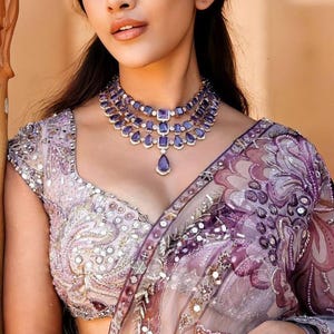 May include: A woman wearing a layered purple gemstone necklace, matching bracelet, and ring. She is wearing a light purple and silver embroidered top and a sheer purple sari with floral patterns. The background is a warm, neutral color.