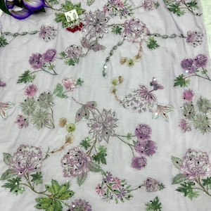 May include: A light purple fabric with embroidered floral patterns in shades of purple, pink, and green. The design includes large flowers, smaller blossoms, and leaves, with scattered sequins for added detail. The fabric is likely used for clothing or home decor.