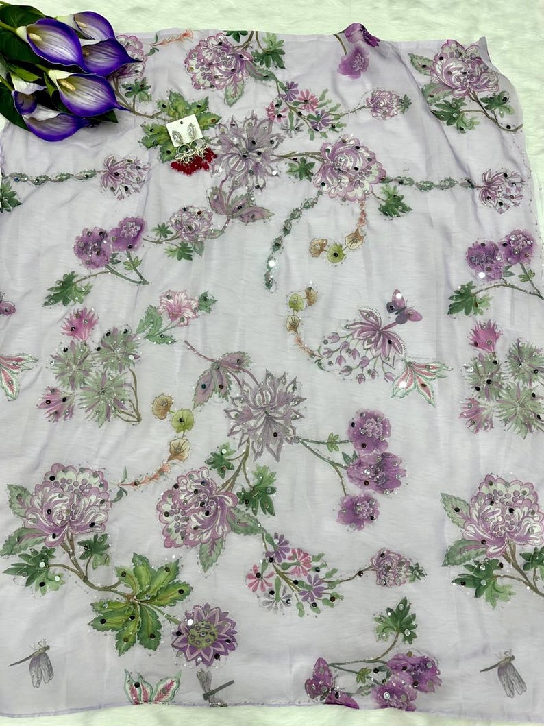 May include: A light purple fabric with embroidered floral patterns in shades of purple, pink, and green. The design includes large flowers, smaller blossoms, and leaves, with scattered sequins for added detail. The fabric is likely used for clothing or home decor.