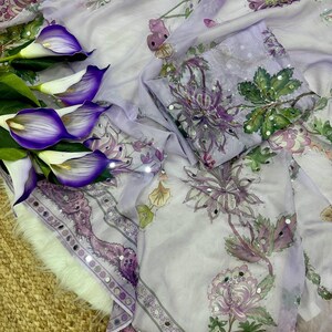 May include: A light purple fabric with floral patterns in shades of purple, green, and white. The fabric has a decorative border with mirrored accents. Purple calla lilies are arranged on the left side of the image.