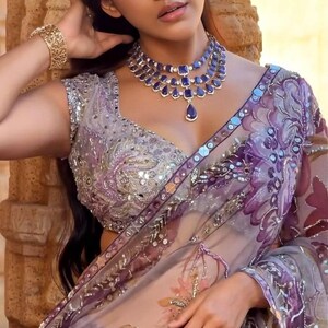 May include: A woman wearing a sheer, lavender sari with floral embroidery and a silver-embroidered blouse. She is adorned with a layered blue gemstone necklace, a matching ring, and a gold bracelet. The sari features purple and pink floral patterns.