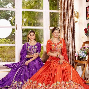 May include: Two women wearing traditional Indian formal wear. One is in a purple outfit, the other in orange. Both outfits feature intricate embroidery with floral and geometric patterns. The outfits include matching tops, skirts, and scarves.