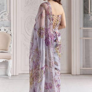May include: A light purple saree with intricate floral embroidery in shades of purple, gold, and green. The saree features a corset-style bodice with lace-up detailing and a sheer, embellished shawl. The fabric is adorned with sequins.