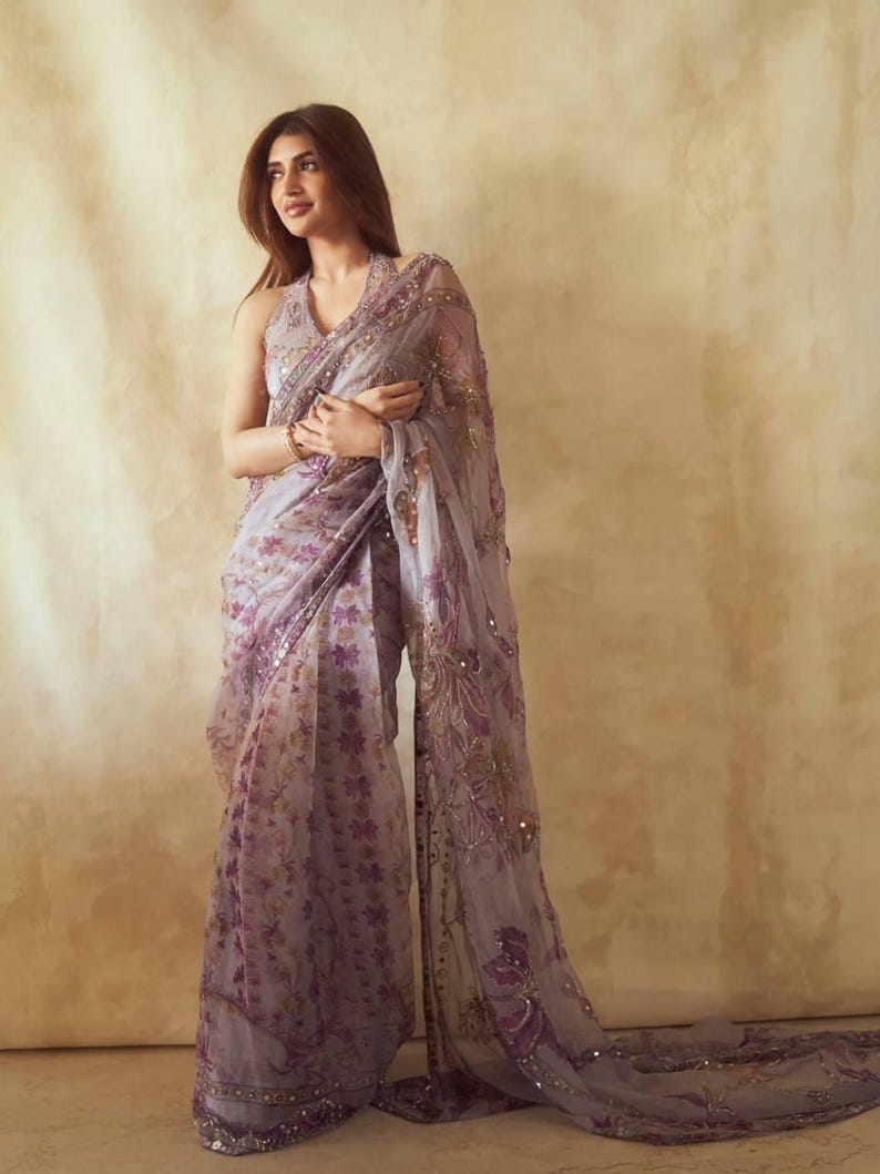 May include: A light purple saree with floral patterns and sequin embellishments. The saree features a halter neckline and a flowing, sheer fabric. The design incorporates shades of purple and gold, creating an elegant and sophisticated look.