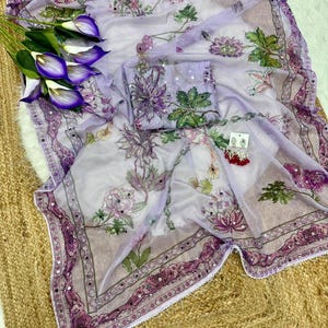 May include: A sheer, lavender-colored fabric with a floral pattern in shades of purple and green. The fabric is adorned with small, reflective embellishments and a decorative border. A matching pouch and earrings are also present.