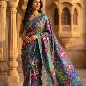 May include: A colorful, patterned saree with a teal border and intricate embroidery. The saree features a patchwork design with shades of blue, green, purple, and pink. The fabric has a shimmering quality, and the border is embellished with small mirrors.
