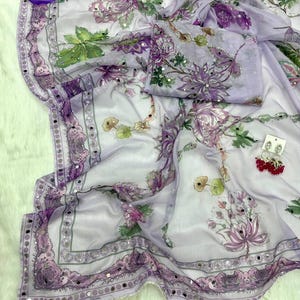 May include: A light purple saree with a floral pattern in shades of purple, green, and white. The saree has a decorative border with small mirrored accents. The fabric is draped, and a matching blouse piece is included.