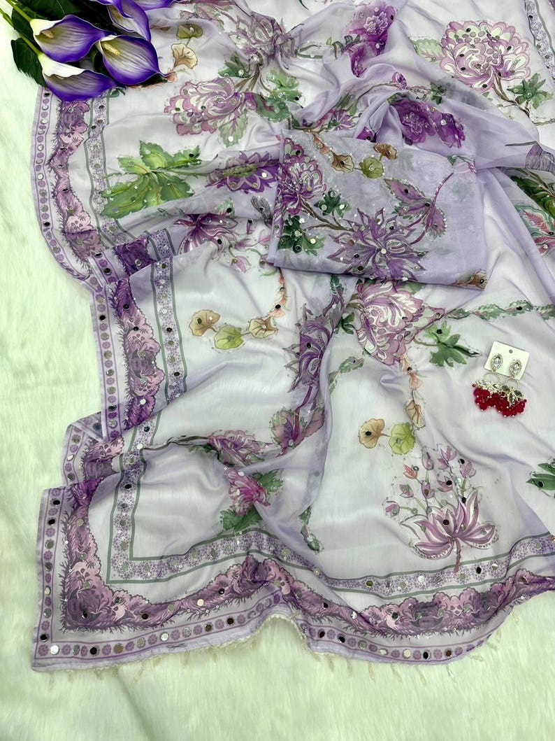 May include: A light purple saree with a floral pattern in shades of purple, green, and white. The saree has a decorative border with small mirrored accents. The fabric is draped, and a matching blouse piece is included.