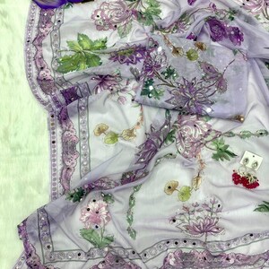 May include: A sheer, lavender-colored fabric with a floral pattern in shades of purple, pink, and green. The fabric is adorned with small, reflective embellishments along the edges and on the floral designs. A matching piece of fabric is folded nearby.