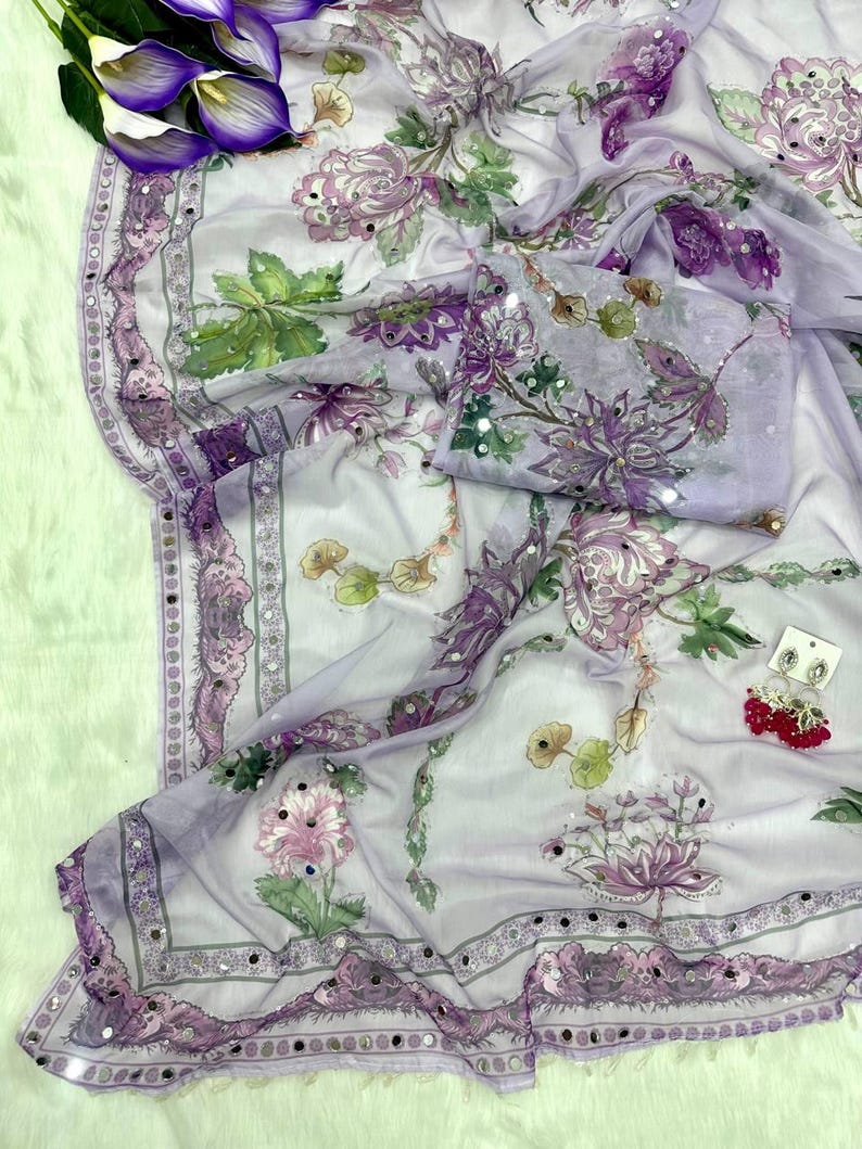 May include: A sheer, lavender-colored fabric with a floral pattern in shades of purple, pink, and green. The fabric is adorned with small, reflective embellishments along the edges and on the floral designs. A matching piece of fabric is folded nearby.