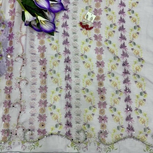 May include: A light purple fabric with a floral pattern in shades of purple, yellow, and green. The fabric has a decorative border with small mirrors and is adorned with artificial purple calla lilies and a pair of earrings.