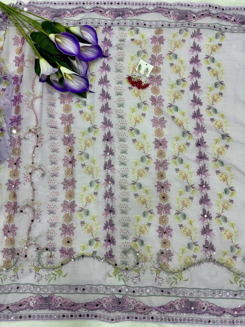 May include: A light purple fabric with a floral pattern in shades of purple, yellow, and green. The fabric has a decorative border with small mirrors and is adorned with artificial purple calla lilies and a pair of earrings.