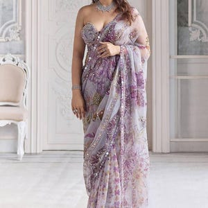 May include: A lavender saree with floral patterns in shades of purple and gold. The saree features a strapless bodice and is adorned with sequins. The fabric is sheer, creating a flowing effect. The overall design is elegant and suitable for formal events.