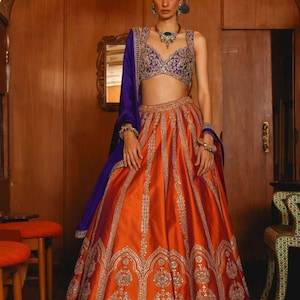 Op de afbeelding: Een vrouw draagt een traditionele Indiase outfit. De set bestaat uit een feloranje rok met ingewikkelde zilveren borduursels, een paars en gouden top en een bijpassende paarse sjaal. De outfit is voorzien van sieraden.