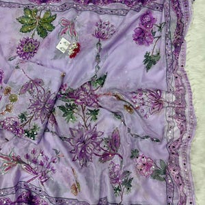 May include: A light purple floral scarf with a matching pouch. The scarf features a repeating pattern of purple flowers and green leaves. The edges are trimmed with a dark purple border and small silver embellishments. The fabric appears to be lightweight and flowing.